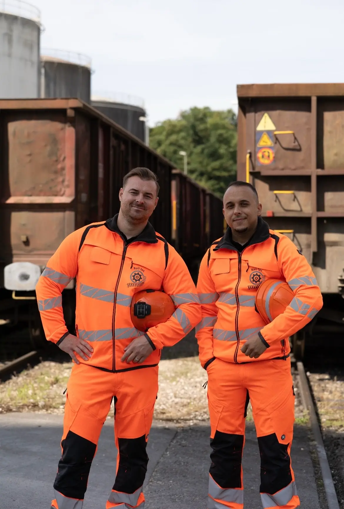 The two Rail-tec specialists in orange high-visibility workwear, helmets under their arms, between two freight wagons on a siding.