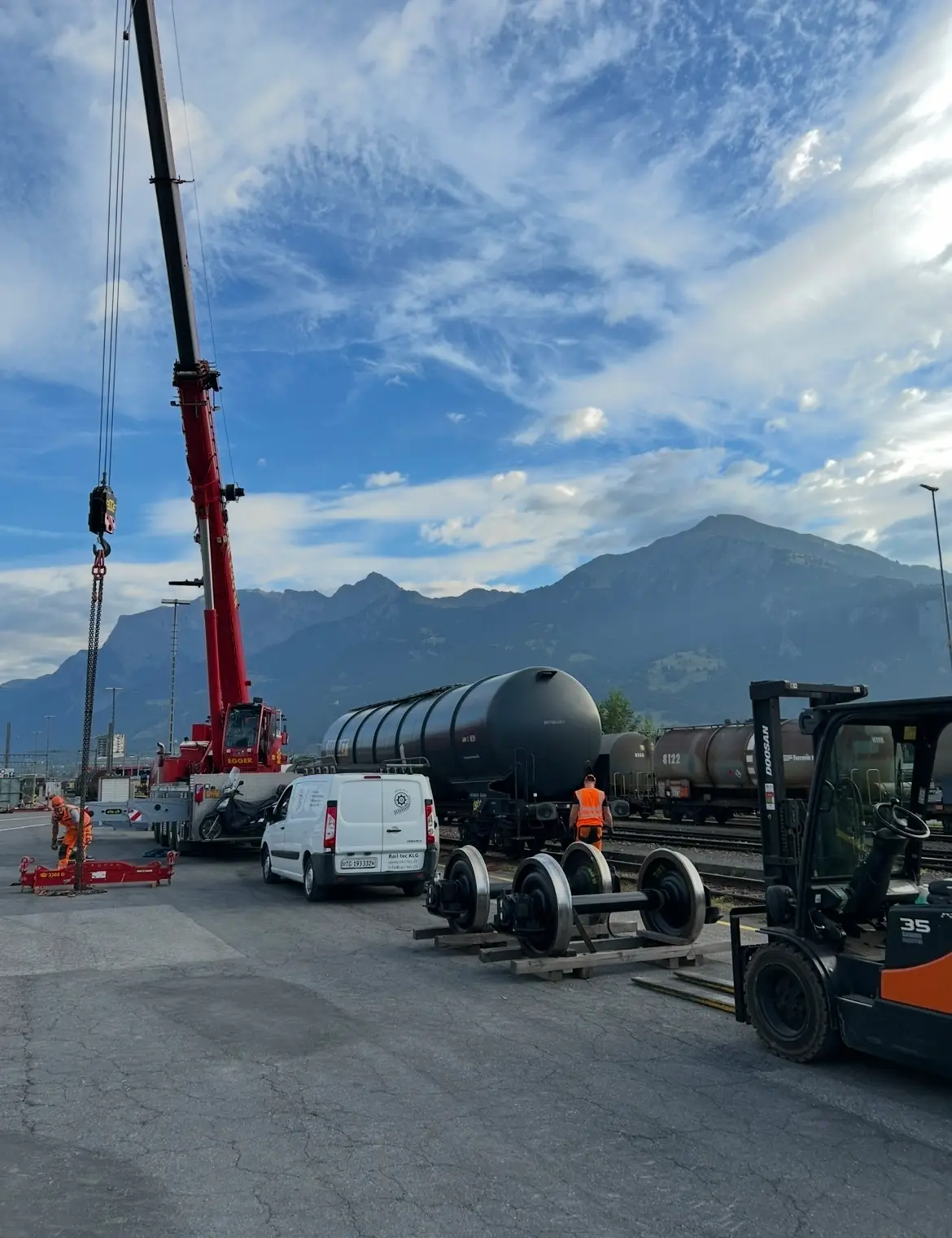A mobile crane over the track field, the Rail-tec service van in front of it, wheelsets set out on sleepers and a forklift; tank wagons and mountains behind.
