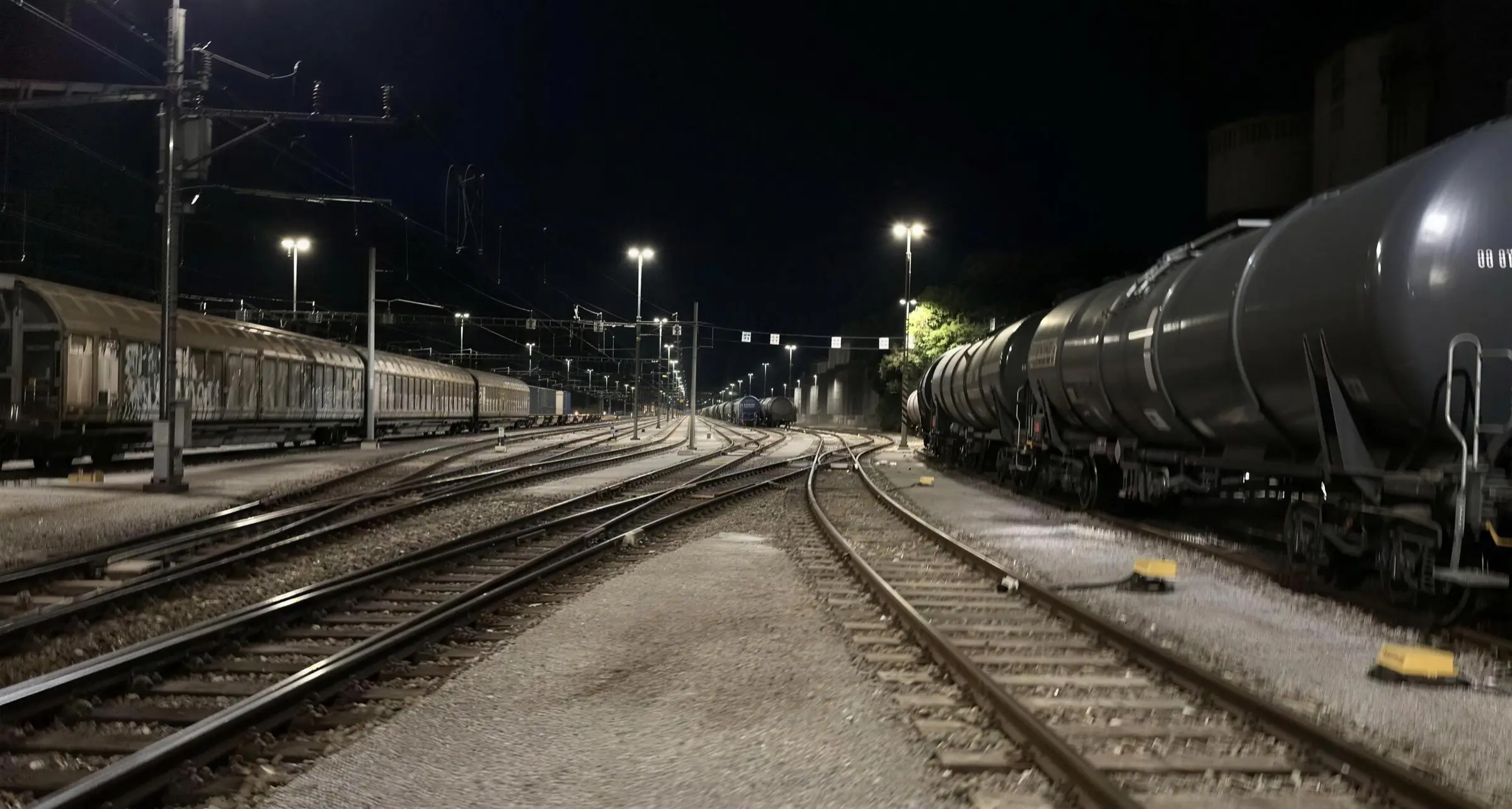 A marshalling yard at night: the tracks converge under the lamps, a line of tank wagons to the right, stabled freight wagons to the left.